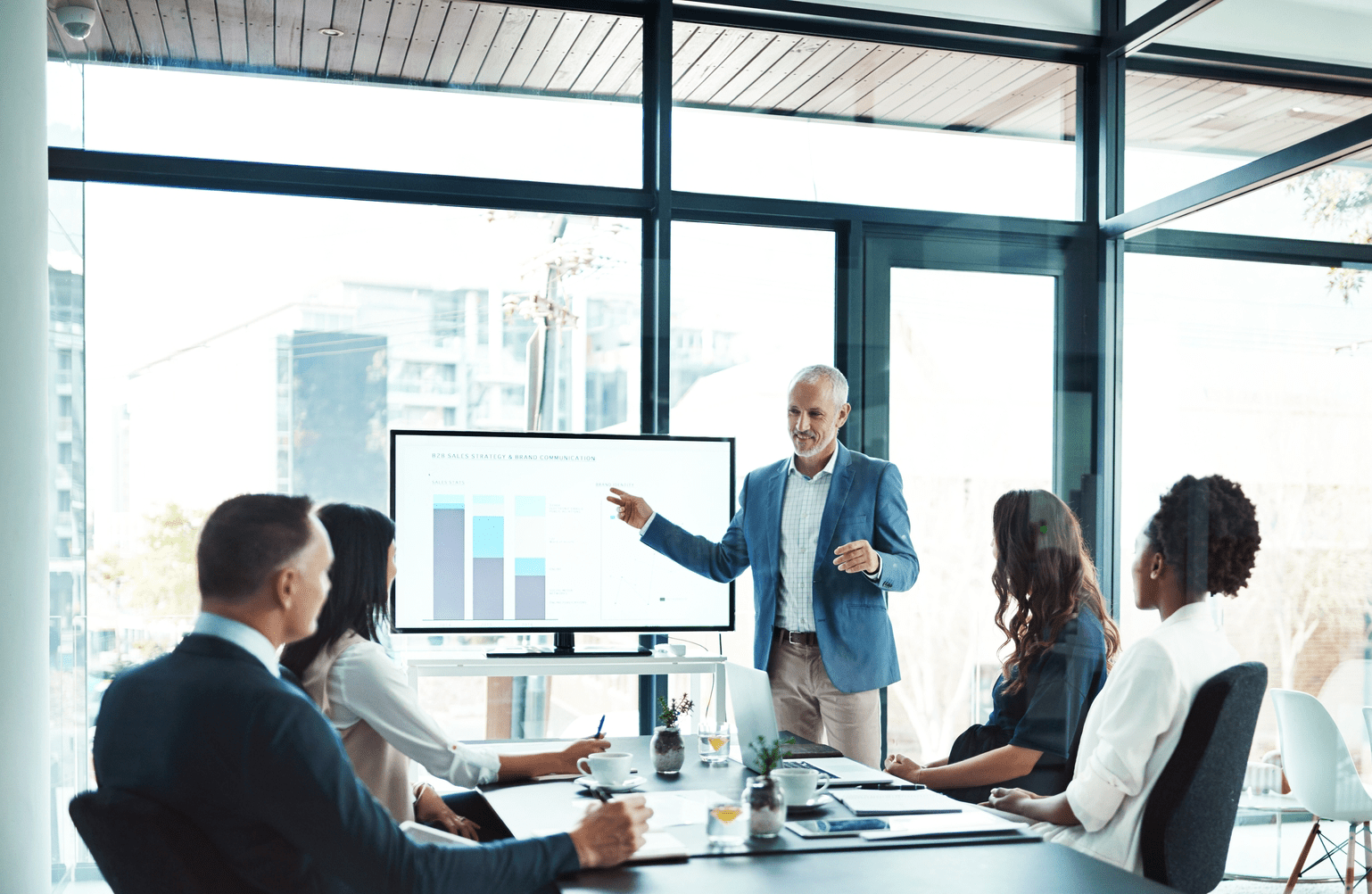 A presenter gestures towards a screen displaying a sales strategy graph, while six colleagues attentively engage in a modern conference room.